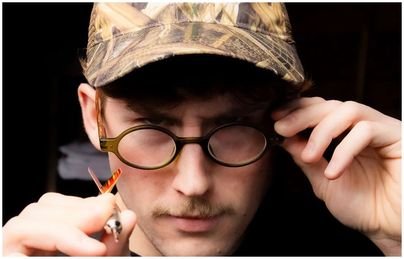 Young man wearing camouflage hat and round glasses, holding a cigarette near his lip, staring intently, dark background.
