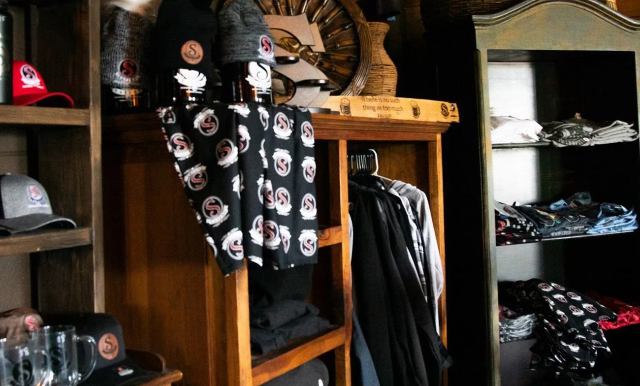 Shelves of hats, shirts, and other apparel, with a wooden sign on top of a clothing rack reading 'there is too much thing as too much'.