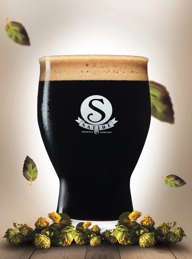 A glass of dark stout beer with a frothy head, branded with the Sature Brewing Company logo, surrounded by hops cones and floating hop leaves on a wooden surface.