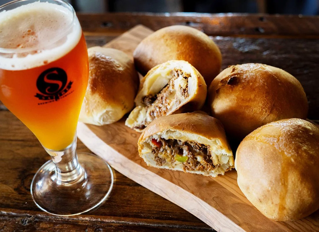 A glass of beer next to a wooden serving board with several baked meat-filled buns, some of which are cut in half showing the filling inside.