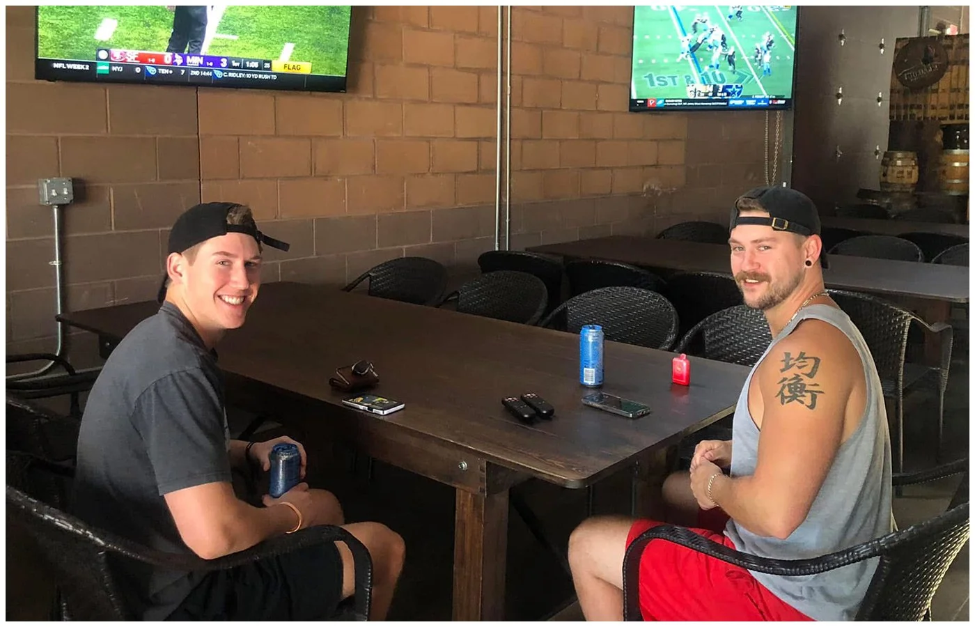 Two men sitting at a wooden table in a bar, smiling and holding cans of soda. Two TV screens showing football games are mounted on a brick wall behind them, and there are empty chairs and tables around them.