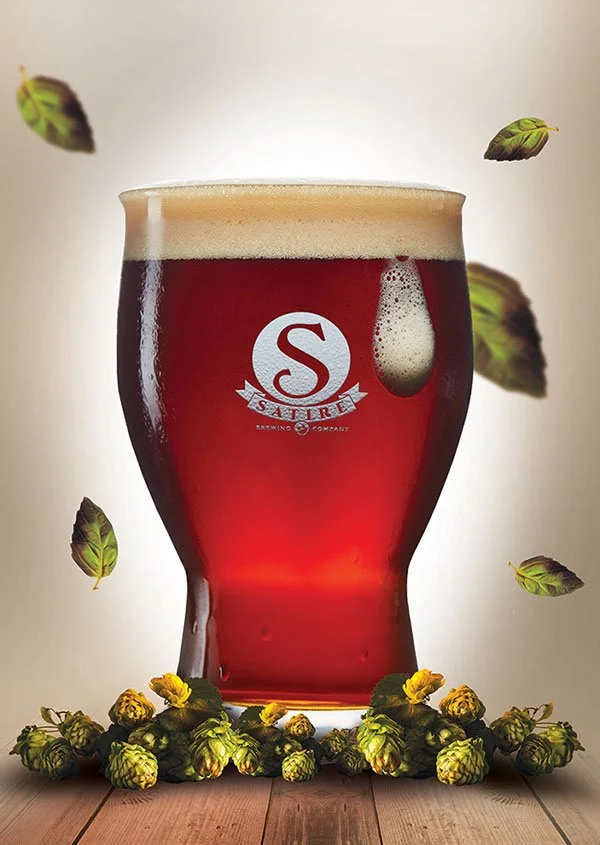 A glass of red beer with a foam head, surrounded by hops and hop cones, with a logo on the glass.