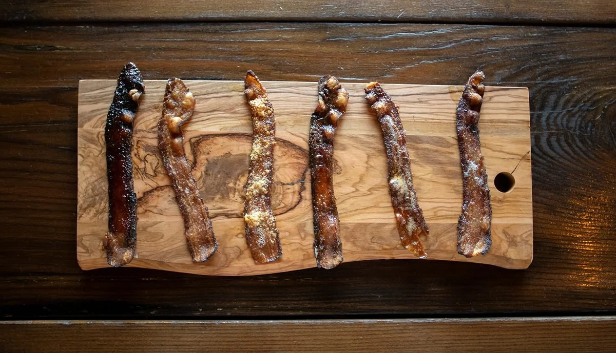 Six bacon strips with sugar on a wooden serving platter on a dark wooden table.