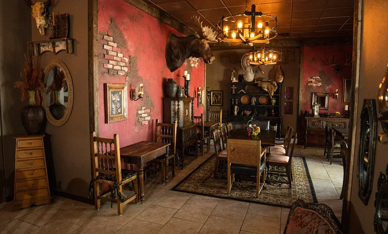 A rustic dining room with a large dark wood table and chairs, decorated with mounted animal heads including a moose and a deer, antique furniture, and a chandelier with candles hanging from a wooden ceiling. The room has exposed brick and distressed walls, with various decorative items and artwork.