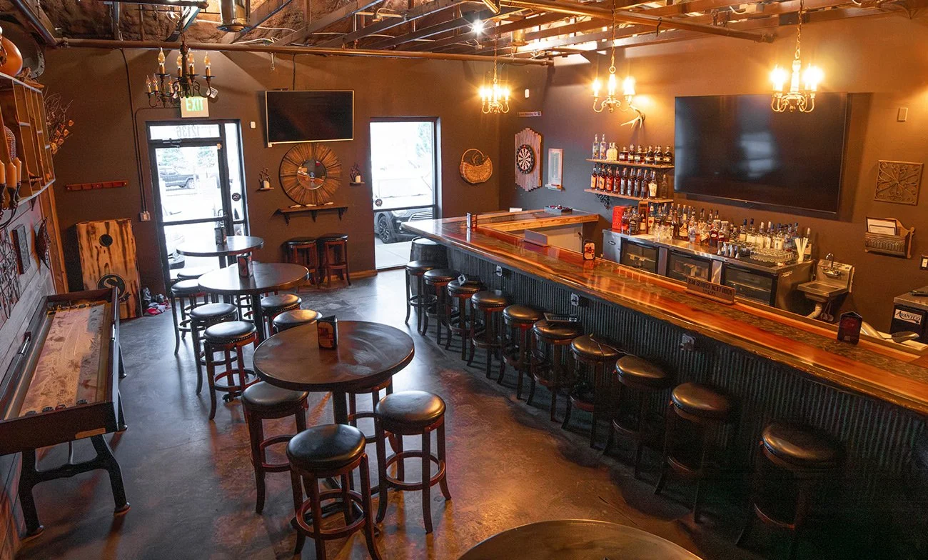 Interior of a bar with a long wooden counter, black barstools, a dartboard on the wall, multiple TV screens, high tables with barstools, warm lighting from chandeliers, and decorative wall art.