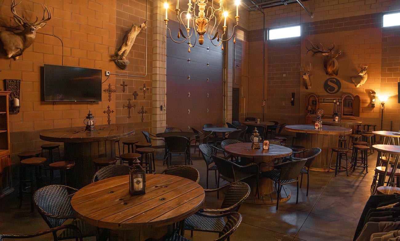 Cozy restaurant interior with brick walls, mounted deer and animal heads, round wooden tables, black chairs, and a chandelier hanging from the ceiling.