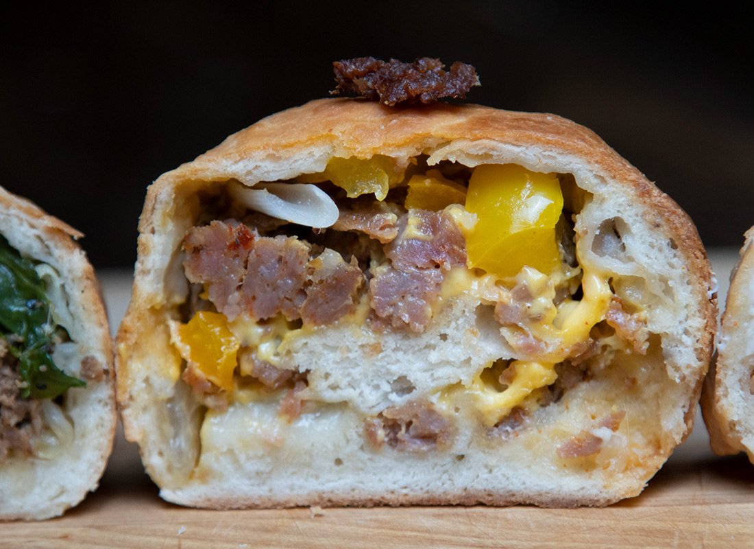Close-up of a baked biscuit filled with scrambled eggs, cooked sausage, and melted cheese, topped with a small piece of cooked bacon, on a wooden surface.