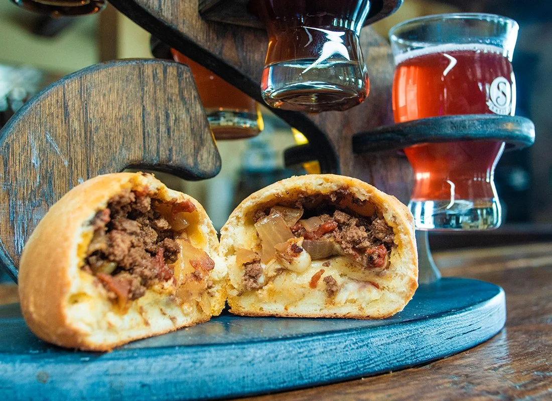A beef pot pie sliced in half, showing ground beef, vegetables, and gravy inside a golden crust, with glasses of beer or cider in the background on a wooden serving tray.