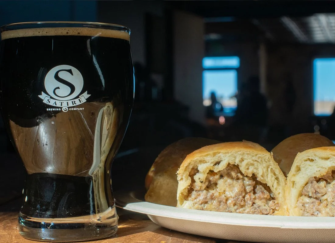A pint glass of Dark Beer from Satire Brewing Company next to a sliced meat pie on a white plate, with a dark indoor background and a window showing a blue sky.