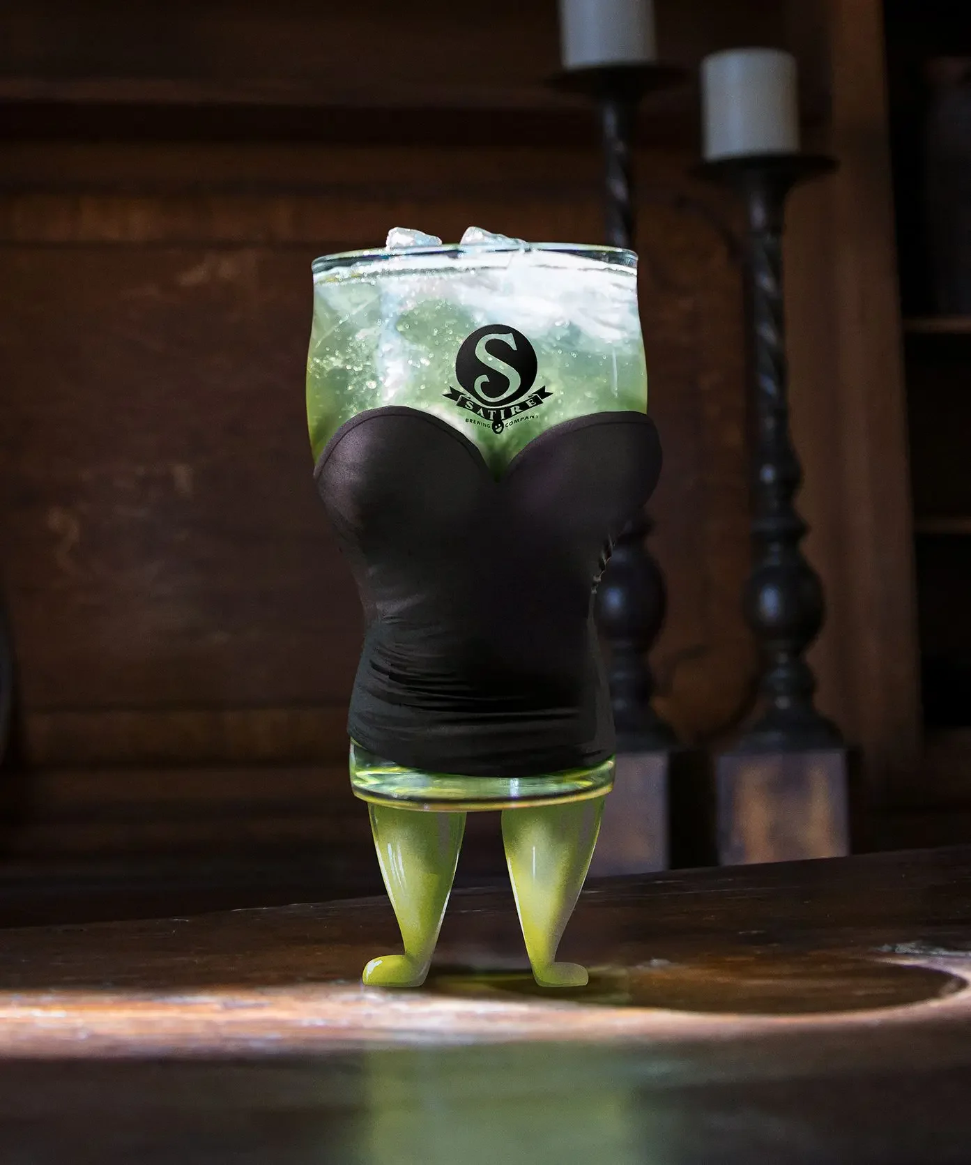A glass of green drink with ice, held by a hand wearing a black glove, with yellow legs and feet, and a logo on the glass that reads 'Satire Brewing Company.'