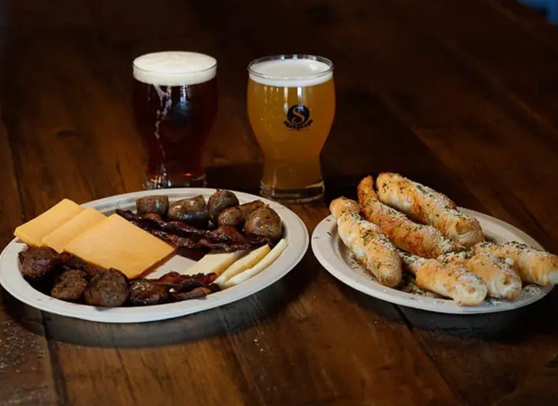 Plate with cheese, bacon, and sausage, and another plate with breadsticks topped with cheese and herbs. Two glasses of beer behind the plates on a wooden table.