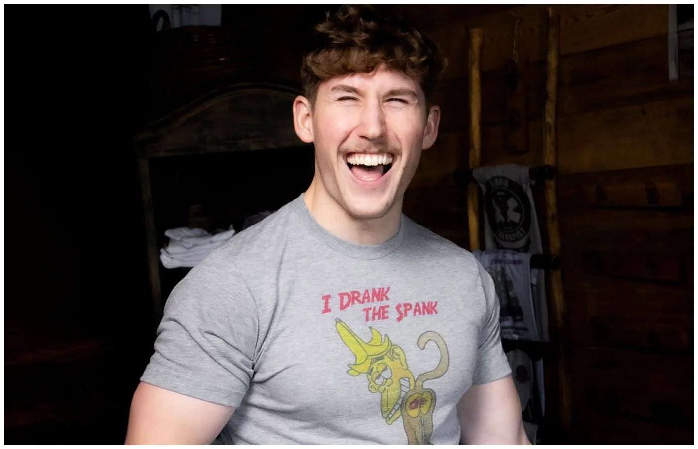 A young man with curly hair and a gray t-shirt with a cartoon banana and the text 'I DRANK THE SPANK,' smiling in a wooden room.
