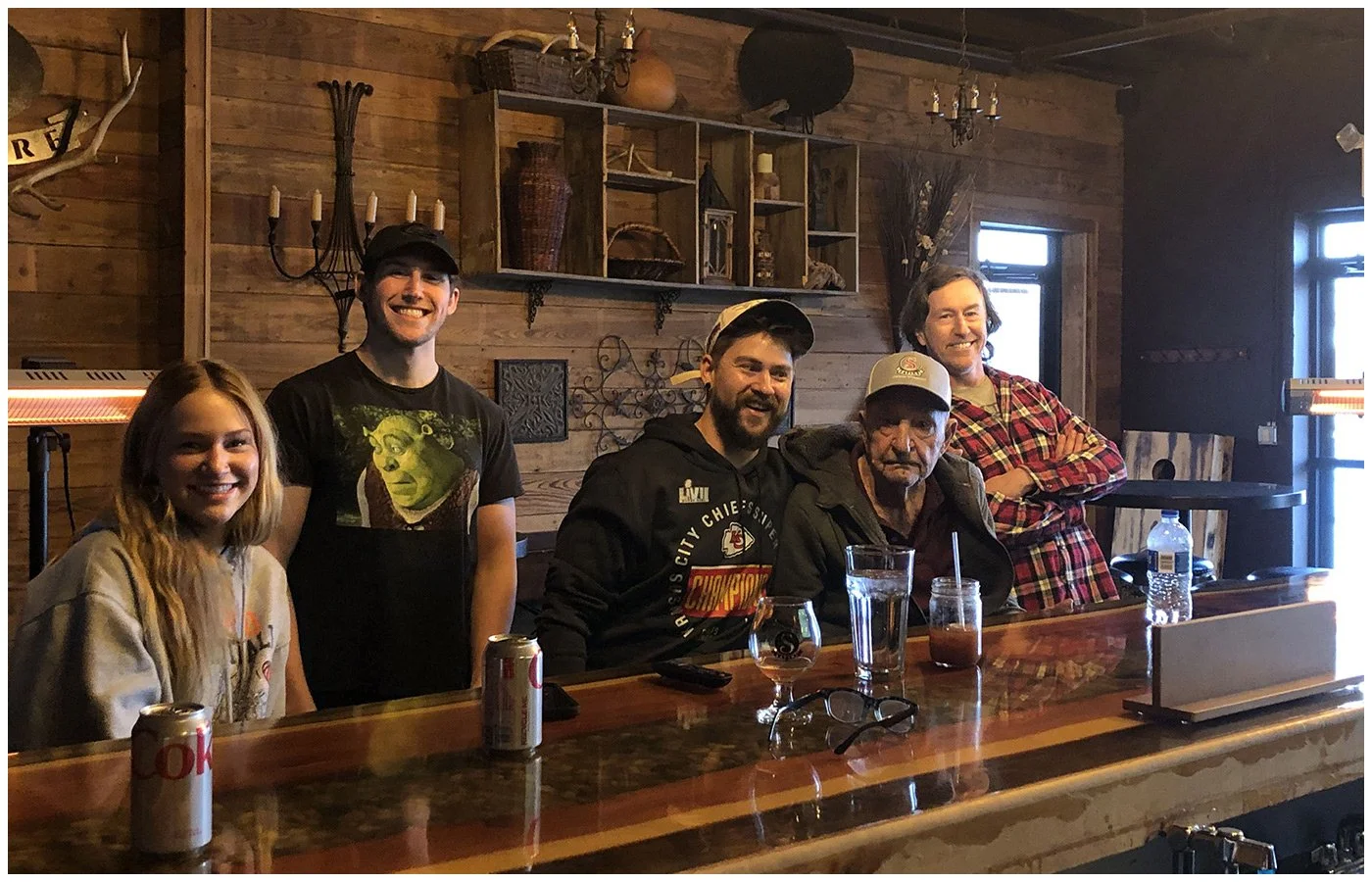 Five people sitting at a bar counter in a rustic wooden interior, smiling, with drinks and cans in front of them.