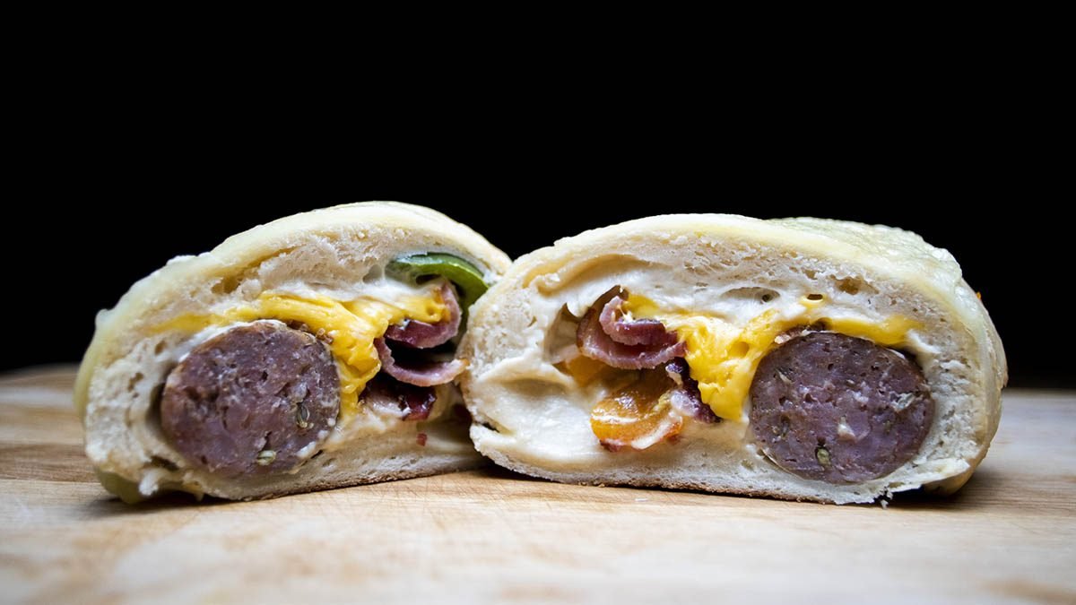 Cross section of a breakfast calzone with sausage, scrambled eggs, cheese, and vegetables on a wooden surface against a black background.