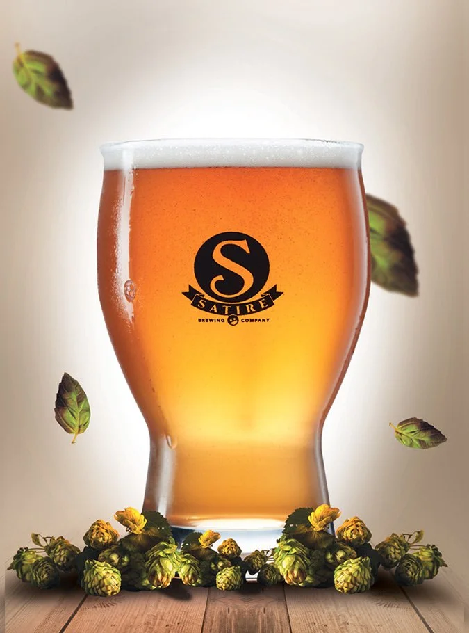 A glass of beer with a logo from Sature Brewing Company, surrounded by hops on a wooden surface with hop pellets and leaves floating around.