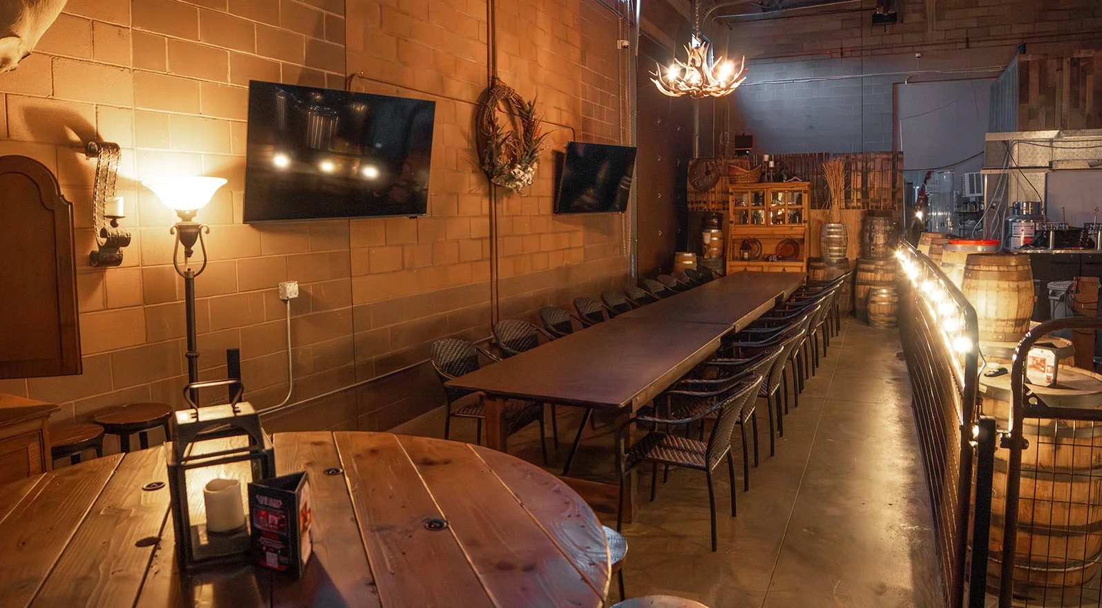 A cozy restaurant or bar with a long wooden table, multiple chairs, and warm ambient lighting. Decor includes mounted TVs, barrel accents, and rustic decor elements.