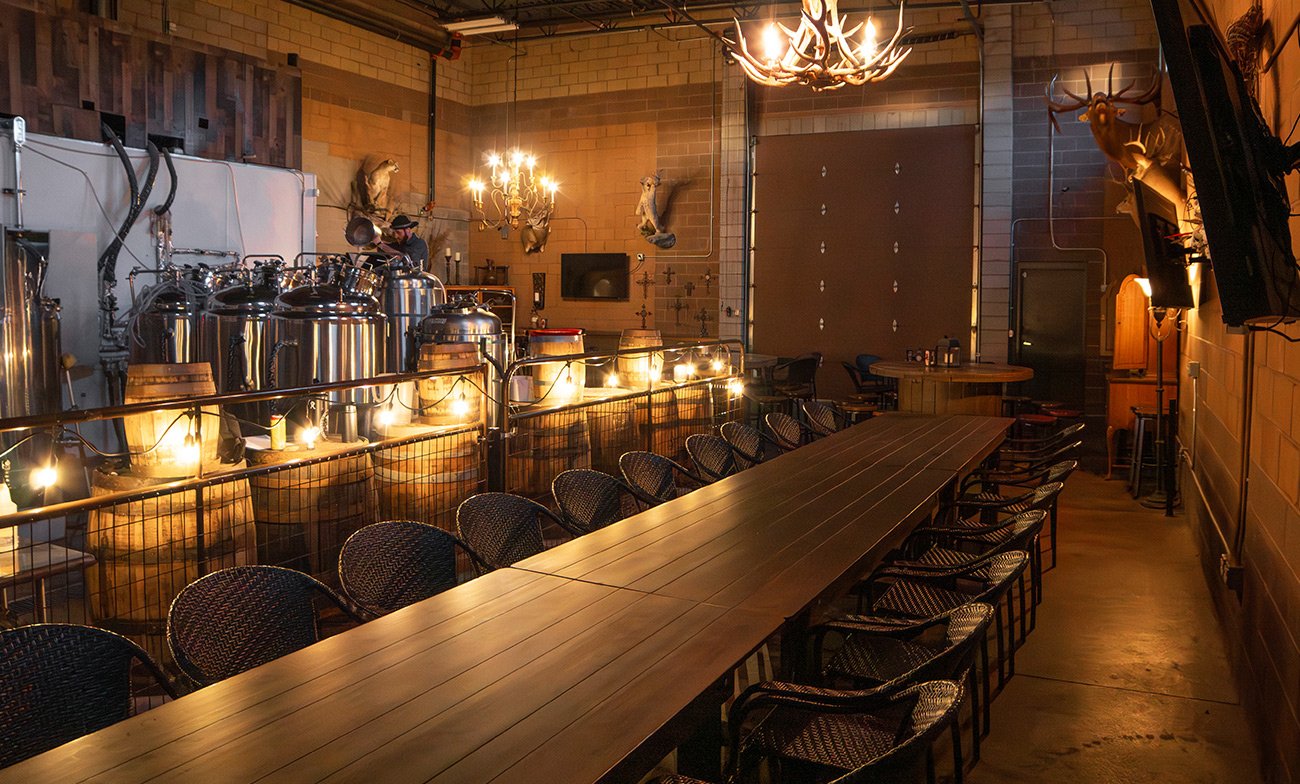 Interior of a rustic brewery or taproom with long wooden table and black woven chairs, chandeliers, mounted animal heads, brewing equipment, and warm lighting.