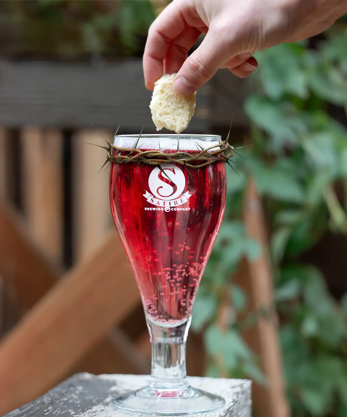 A hand holding a piece of bread over a glass of red beverage with a crown of thorns around the rim, with a blurred green outdoor background.