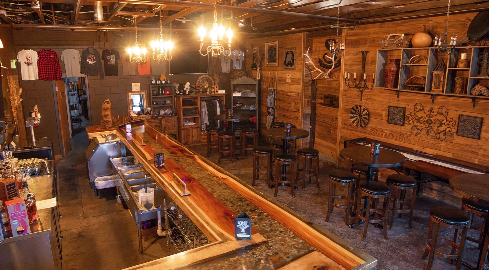 Interior of a rustic bar or pub with wooden walls and furnishings, a long wooden bar counter with barstools, hanging t-shirts on the wall, antique-style lighting fixtures, and decorative items like antlers and artwork.