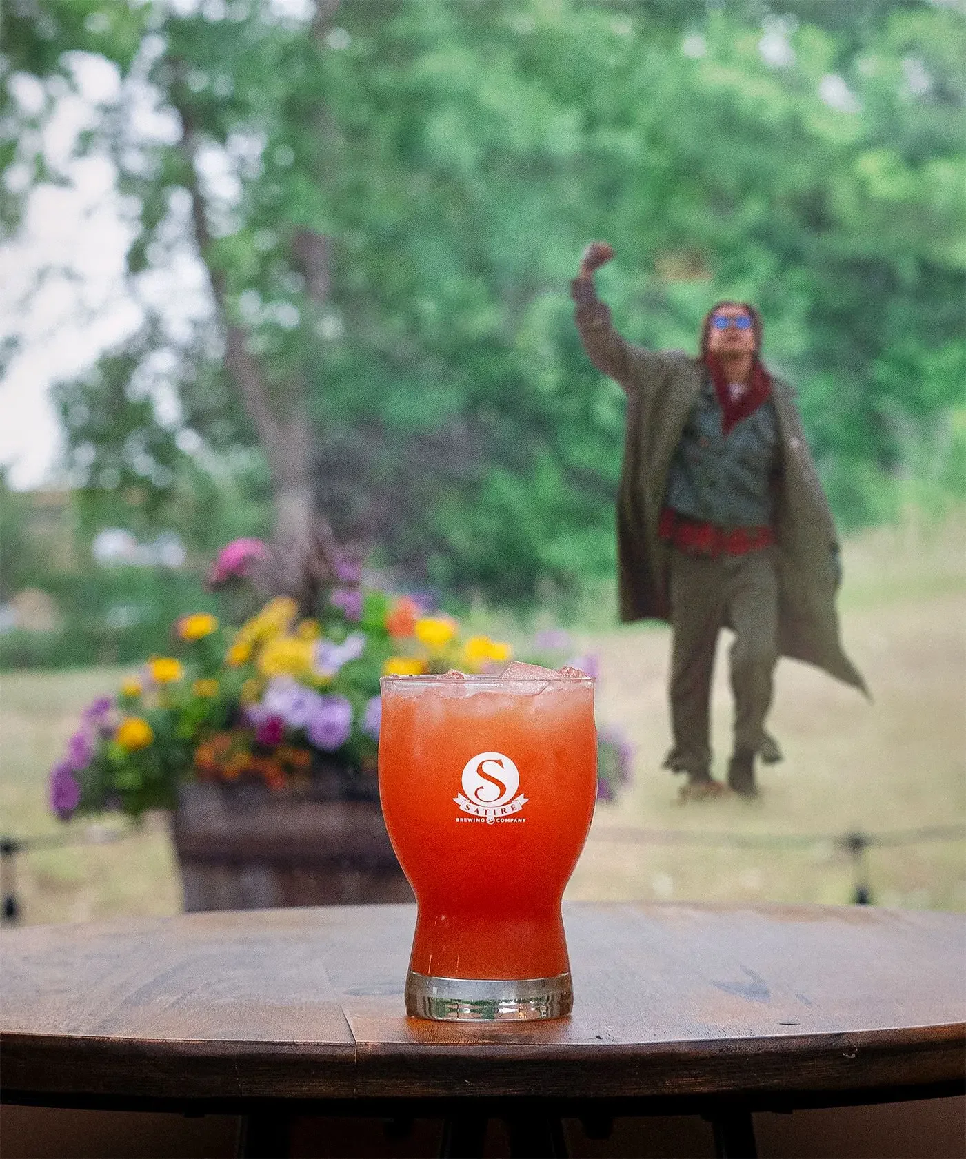 A glass of reddish-orange cocktail with ice on a wooden table. In the background, a man with glasses, red bandana, and brown jacket is walking outdoors near a flower bed and trees, with a blurred image of him raised one arm.