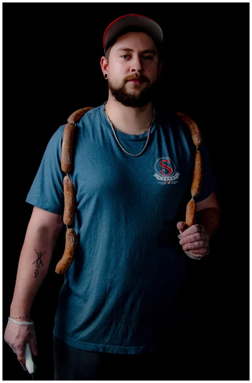 A young man with a beard and black earrings, wearing a baseball cap, a blue t-shirt, and silver necklaces, stands against a black background with a string of sausages draped over his shoulders. He is holding a lighter in his right hand and has a tattoo of scissors and a