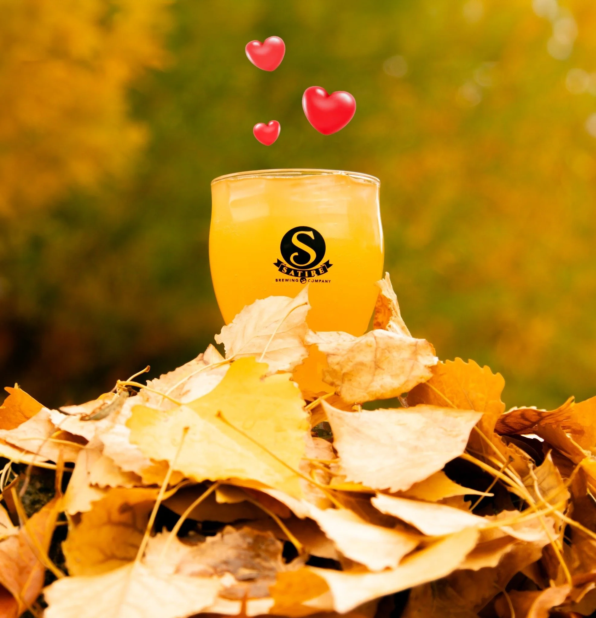 Autumn leaves on the ground with a glass of beer and floating red hearts above it.