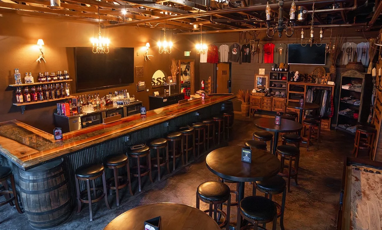 Interior of a rustic bar with wooden furniture, shelves of liquor bottles, a large flat-screen TV, and decorative clothing hanging on the wall.