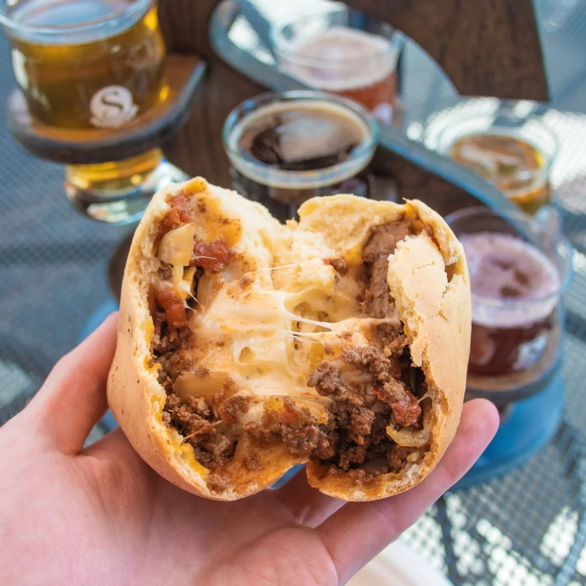 A hand holding a stuffed baked potato with melted cheese, beef, and onions. In the background, there are several small cups of dipping sauces and a glass of beer on a metal outdoor table.