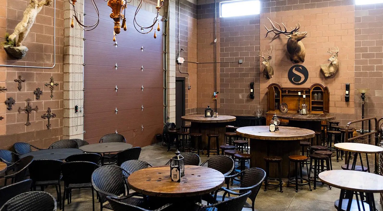 Interior of a rustic-style bar or restaurant with wooden tables and chairs, mounted deer heads, crosses, and a large decorative S on the wall, with a high ceiling and brick walls.