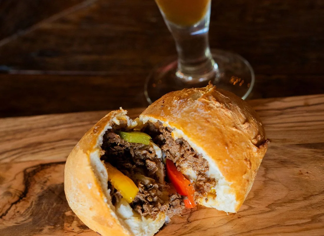 A baked bread roll stuffed with ground beef, bell peppers, and cheese, partially bitten. In the background, a glass of beer on a dark wooden table.