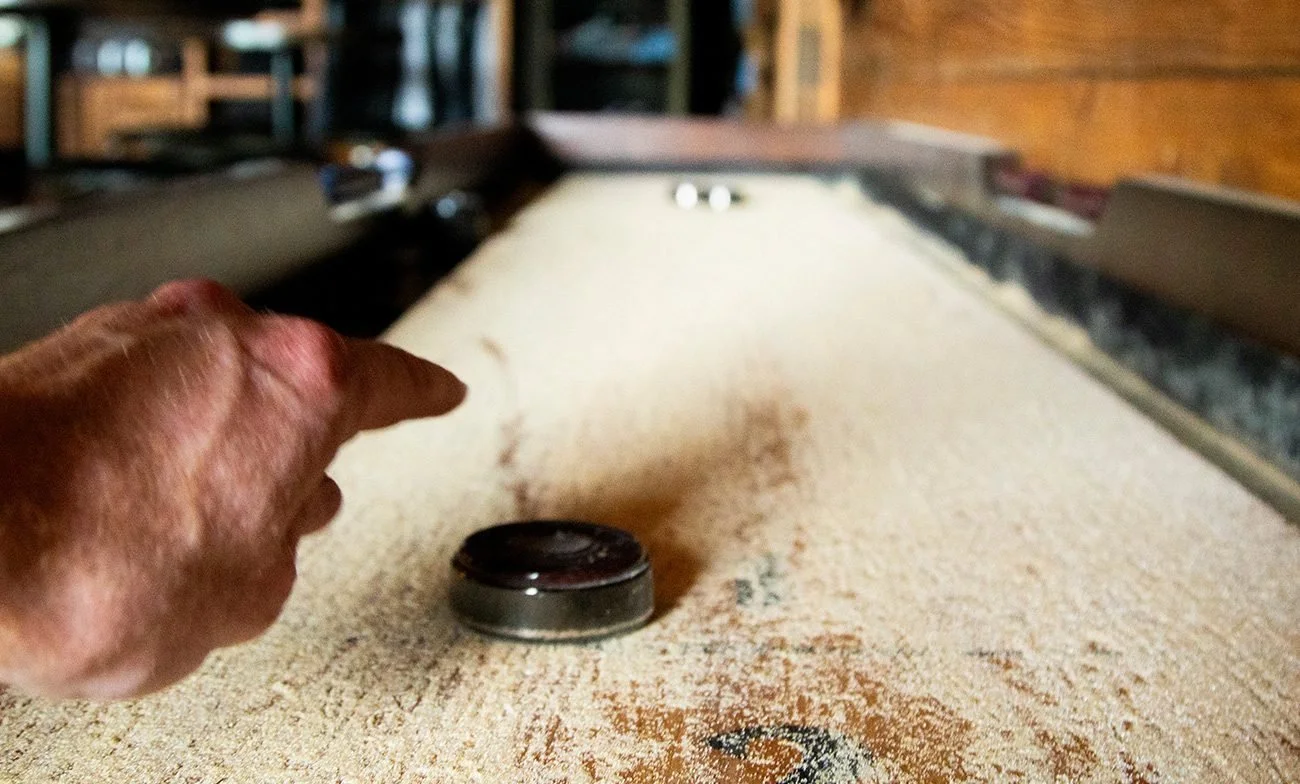 A person is using a hockey puck on a shuffleboard table to slide towards the scoring area. The table is covered with sand or powder, and the hand is pointing or aiming the puck.