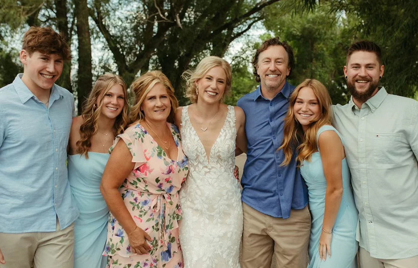 A group of seven people, including a bride in a white wedding dress, standing outdoors in a park with trees in the background, smiling for a photo.