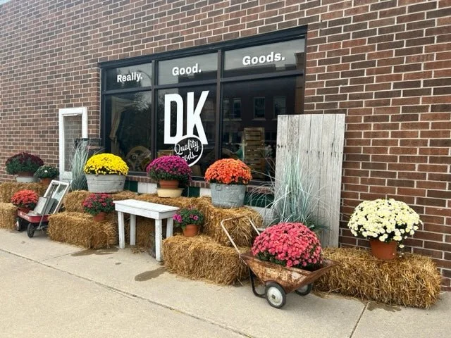 Display of potted chrysanthemums and fall decorations outside a storefront with the sign 'Really. Good. Goods.' and 'DK Quality' in the window.