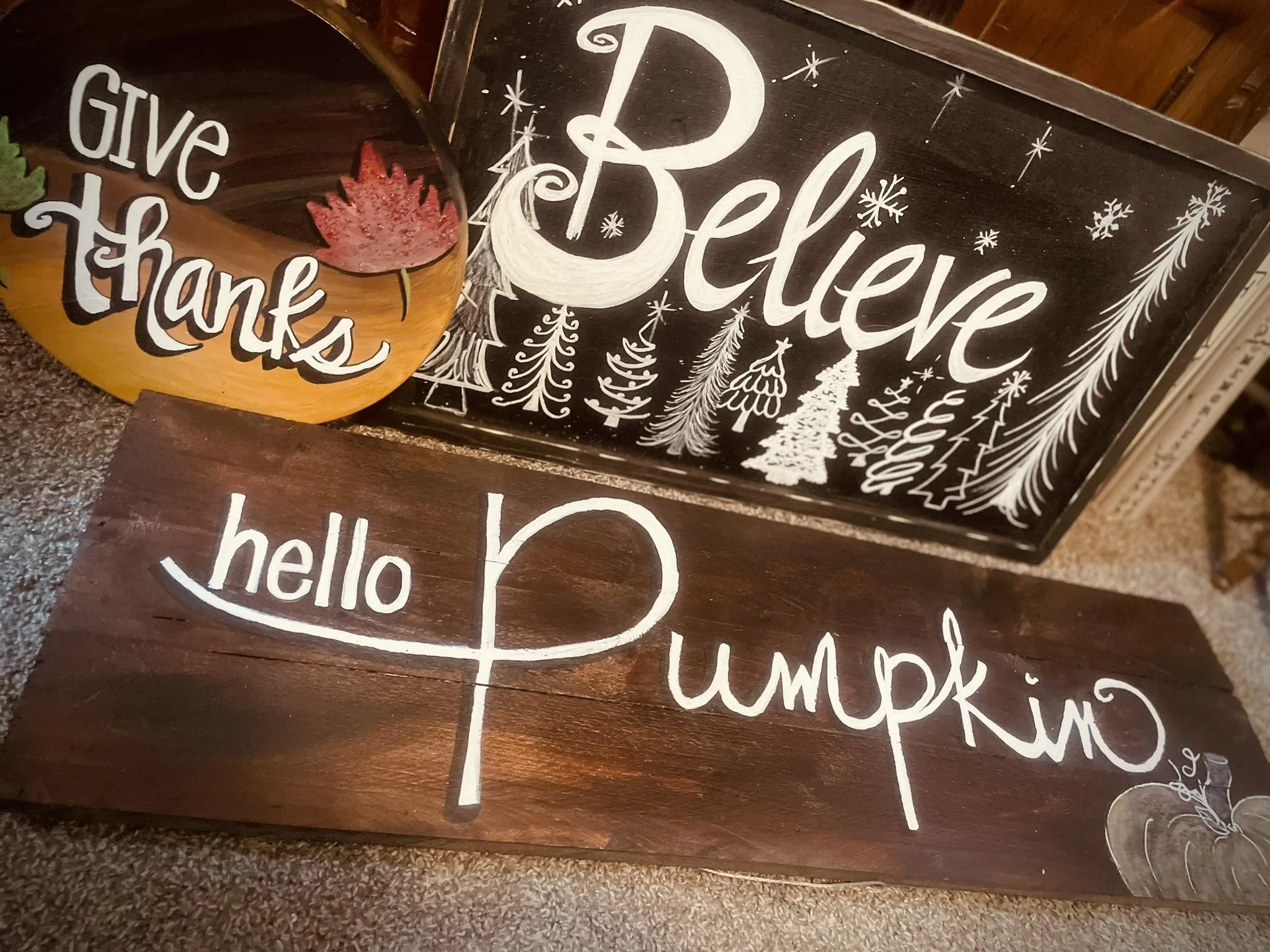 Decorative wooden signs with holiday greeting messages: "Believe" in white with drawings of Christmas trees and snowflakes; another sign says "hello pumpkin" in white with a small pumpkin illustration; an oval sign says "Give Thanks" with a red maple leaf decoration.