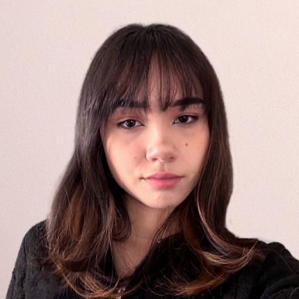 A young woman with straight, dark brown hair and bangs, wearing a black top, posing against a plain light-colored background.