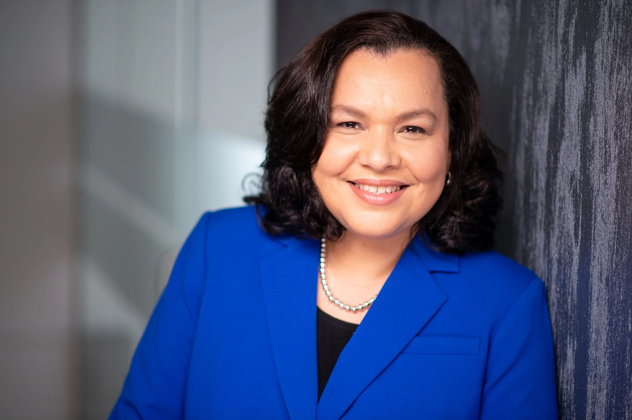 A smiling woman with shoulder-length dark brown hair, wearing a blue blazer, black top, and a pearl necklace, standing against a textured dark wall.