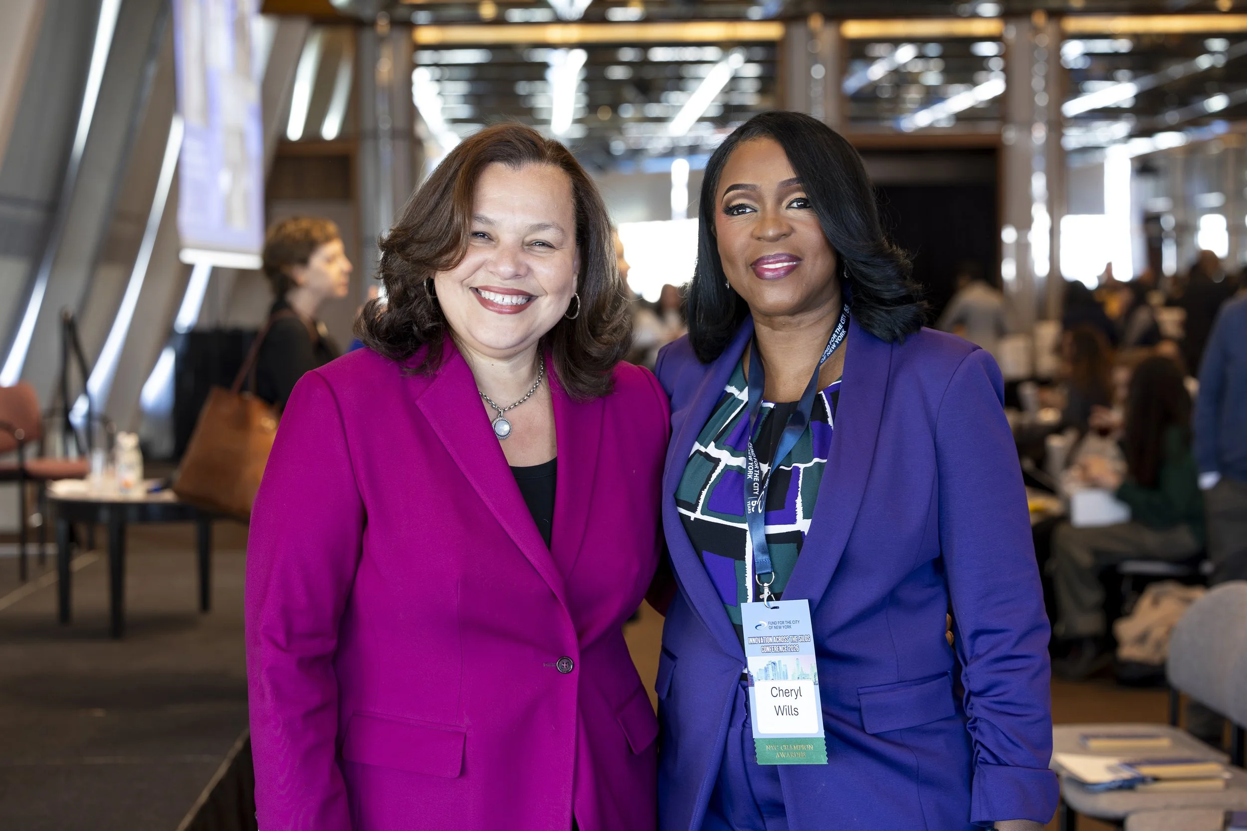 Two women dressed in bright blazer suits smiling at the camera at a conference, with one woman wearing a name badge that says 'Cheryl Wills'.