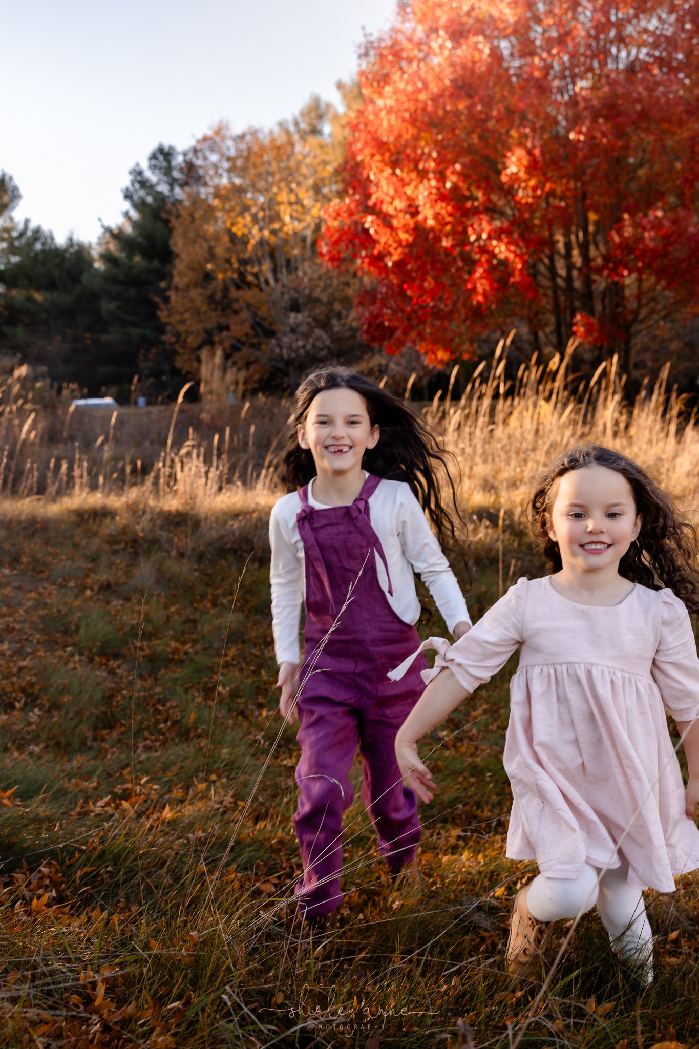 Two young girls running through an autumn field with colorful trees in the background.