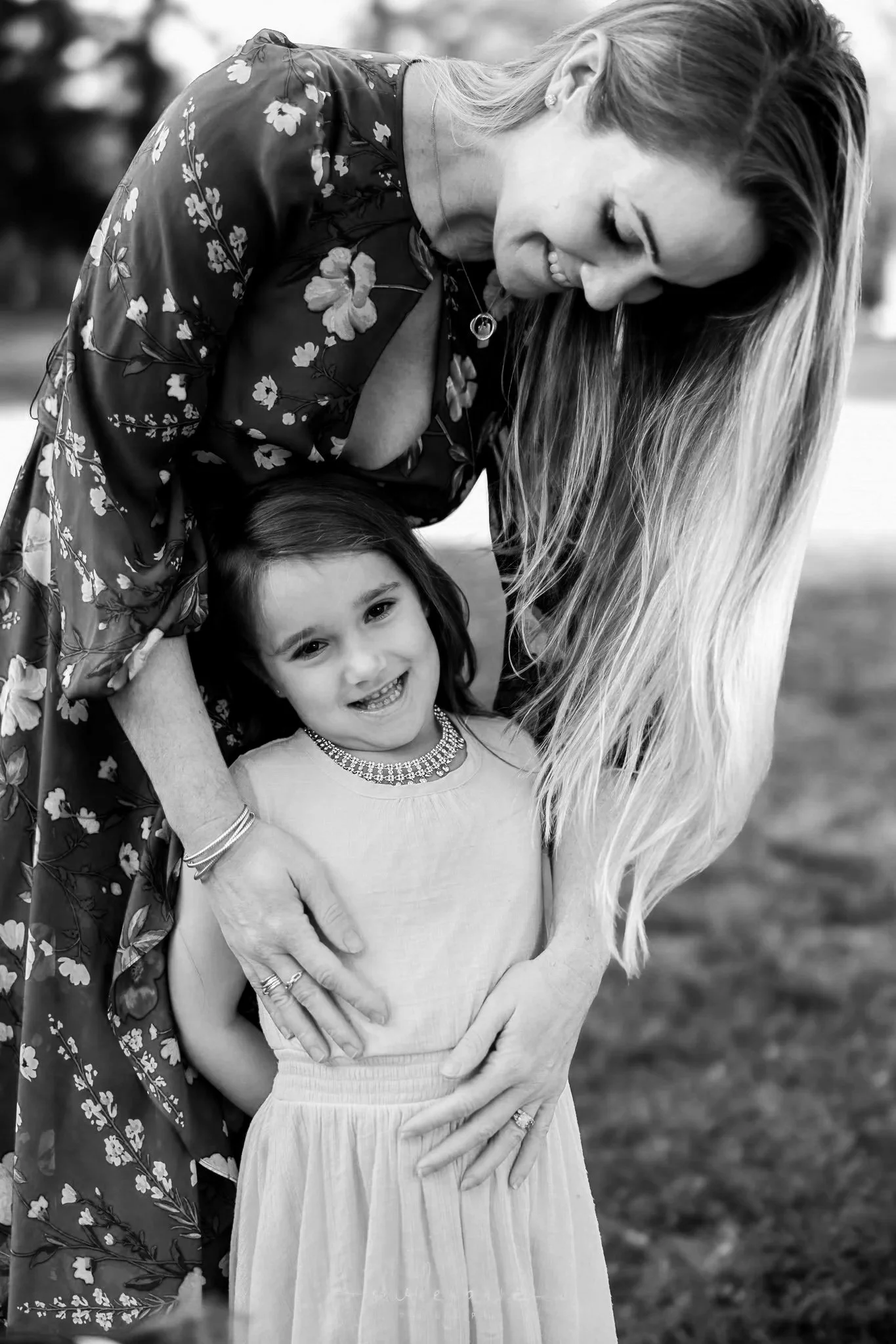 A woman and a young girl smiling outdoors, with the woman leaning over the girl, both looking happy, in a black and white photo.