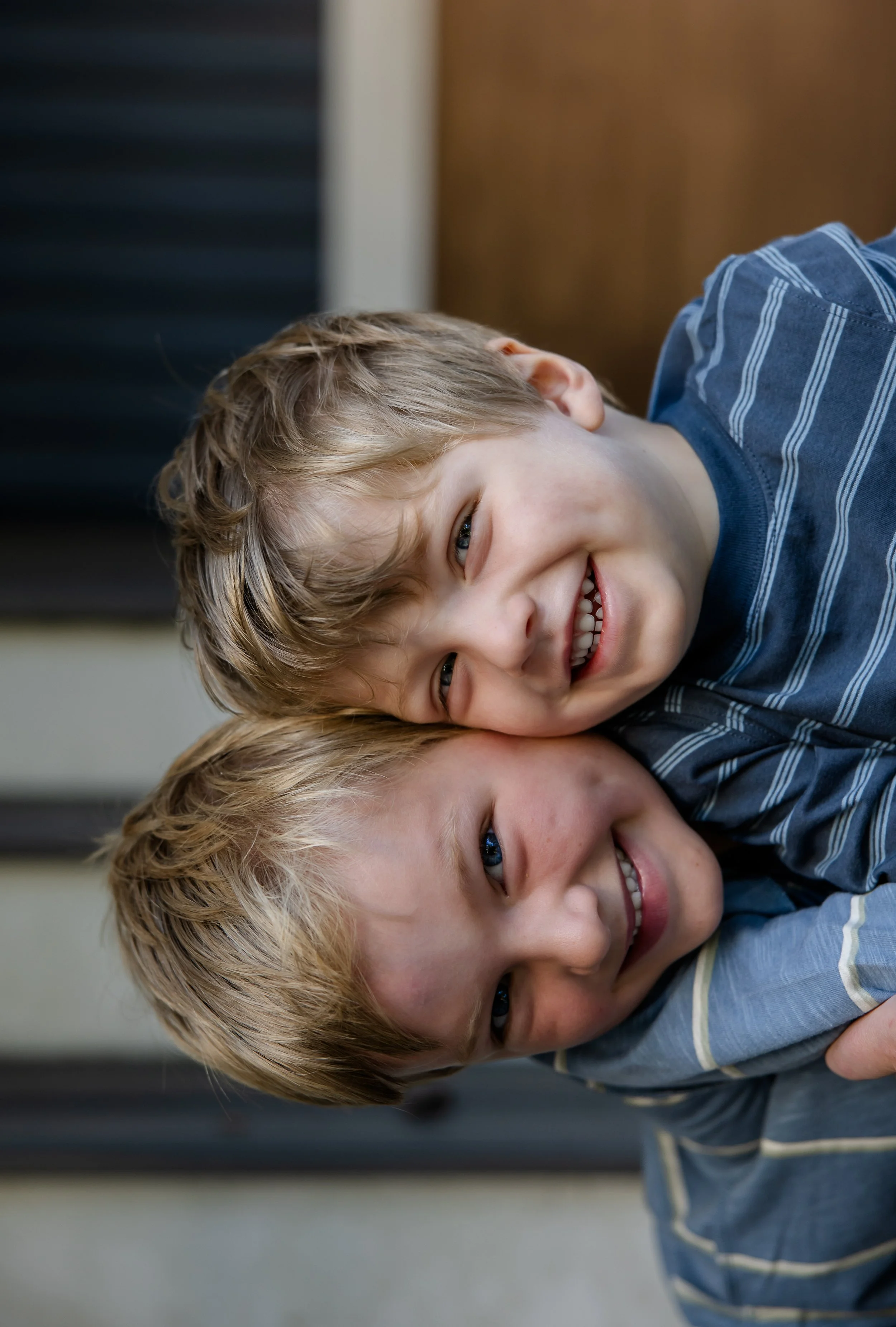 Two young boys with light brown hair smiling and hugging indoors.