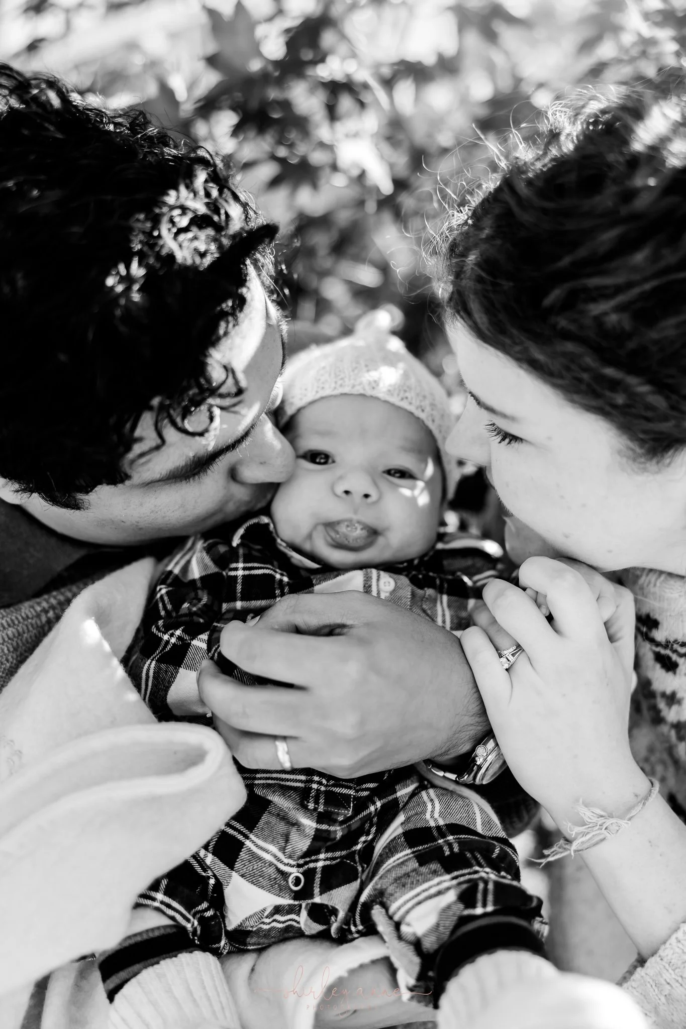 A black and white photo of two adults kissing a baby on each cheek, with the baby in the center looking at the camera and making a facial expression with its tongue out.
