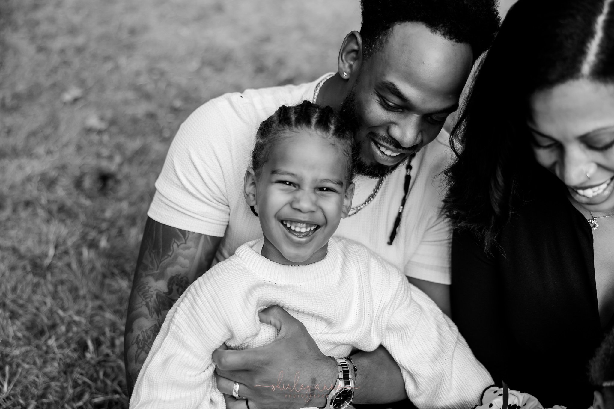 A black and white photo of a smiling young girl with braided hair being hugged by a man and woman, all appearing happy and enjoying a moment outside.