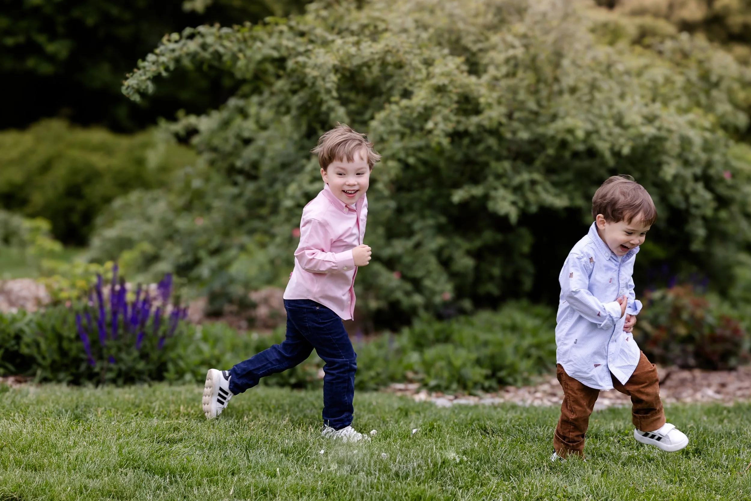 Two young boys playing and running on a lush green lawn with trees and bushes in the background, smiling and enjoying outdoors.