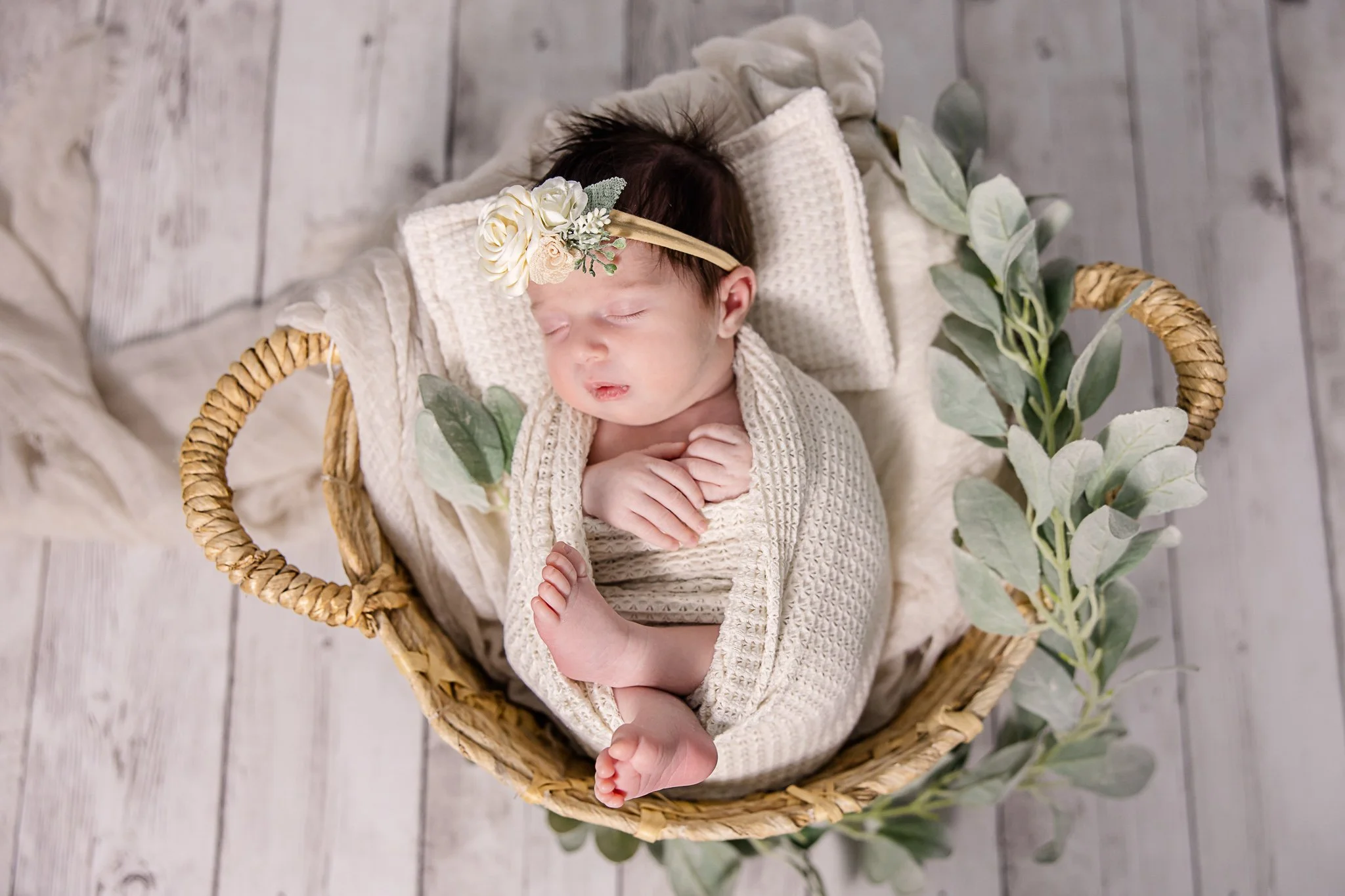 A newborn baby girl sleeping in a wicker basket, wrapped in cream-colored knit blankets, wearing a floral headband with white roses and greenery, surrounded by eucalyptus leaves on a rustic wooden floor.