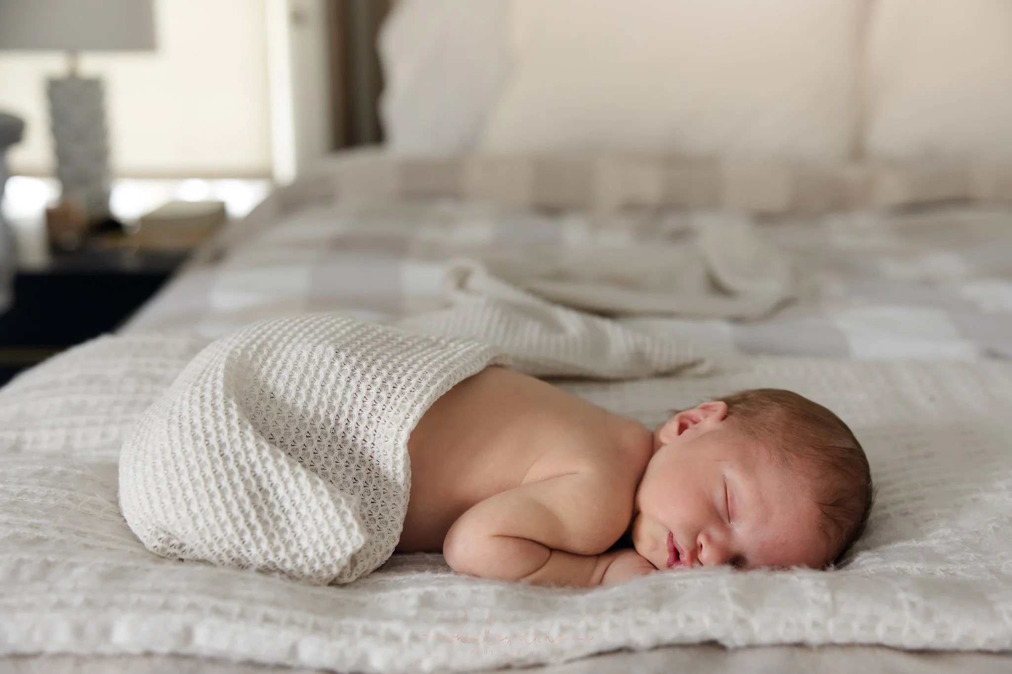 A newborn baby sleeping on a bed, partially covered with a textured white blanket, with a background of pillows and a bedside table.