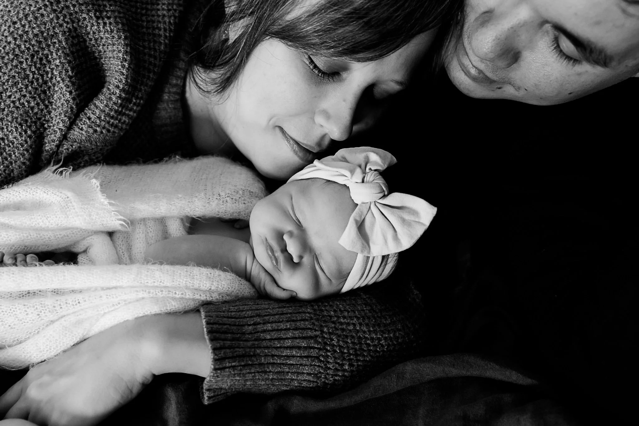 A black-and-white photo of a newborn baby sleeping on a mother's chest, with the mother gently cradling the baby and a man close by.