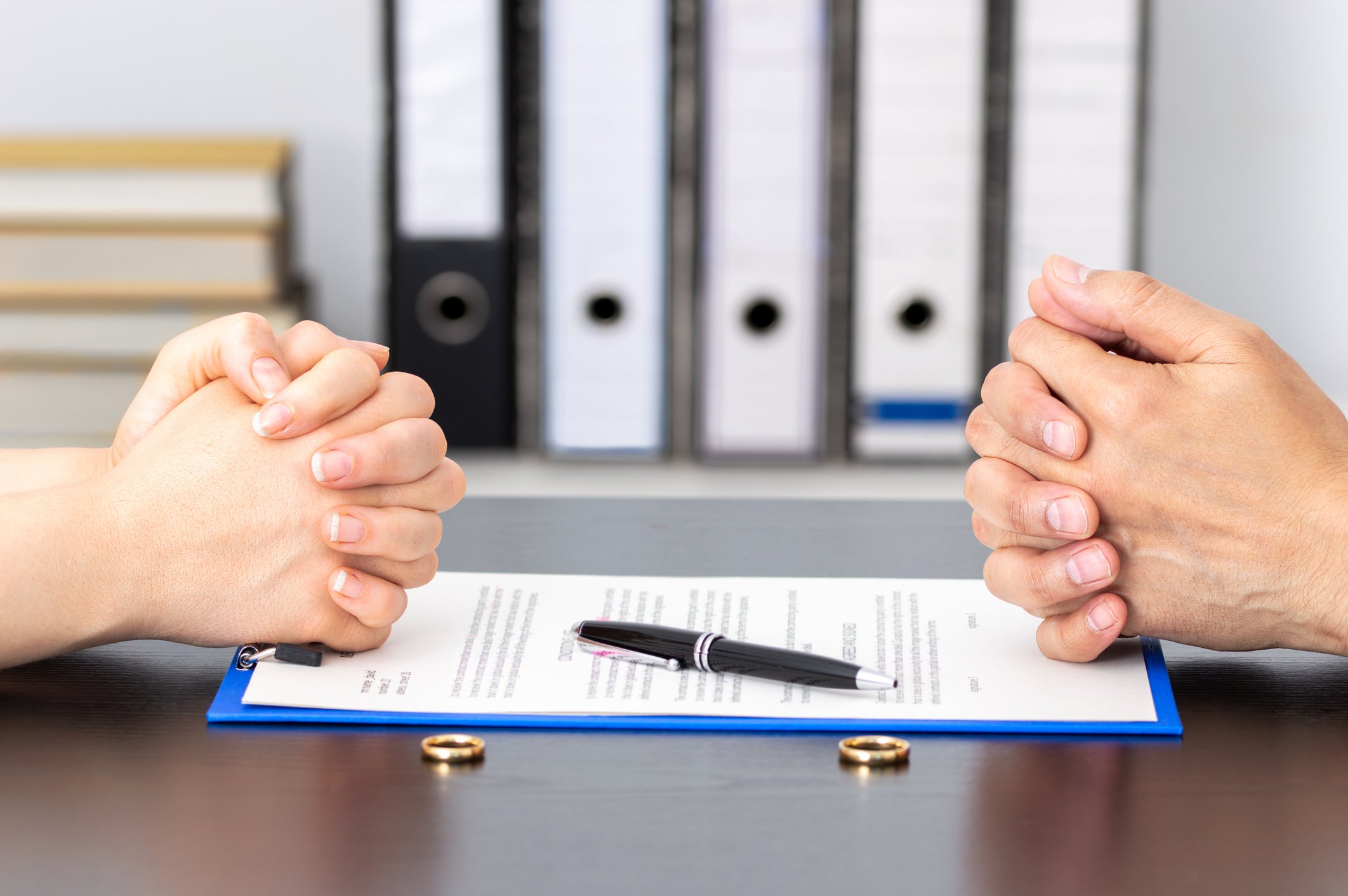 Two people sitting at a table with hands clasped in front of a document, pen, and wedding rings, with office binders in the background.
