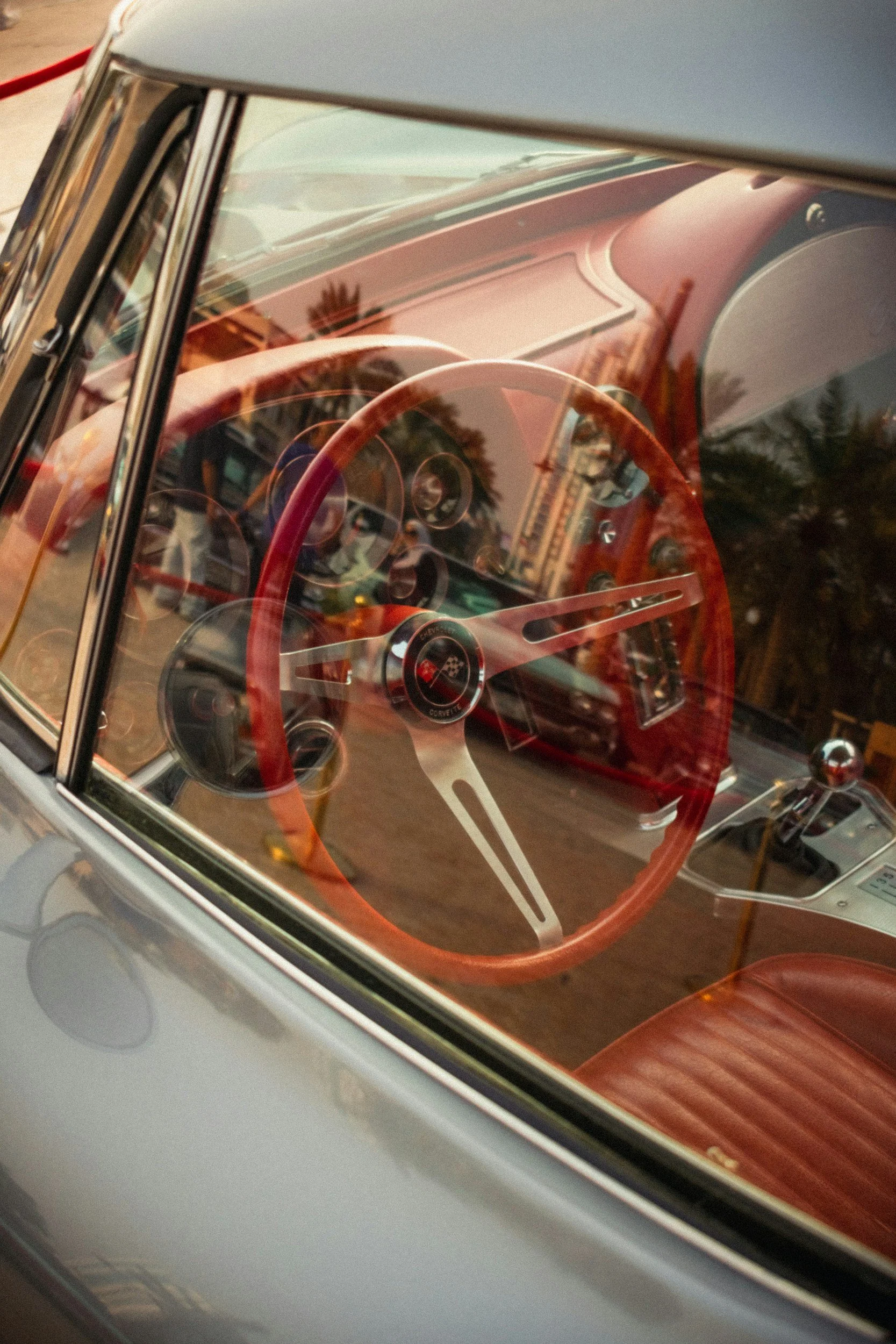 Close-up of a vintage car's interior, showing a red steering wheel, dashboard, and gear shift, with reflections of buildings and trees on the windshield.