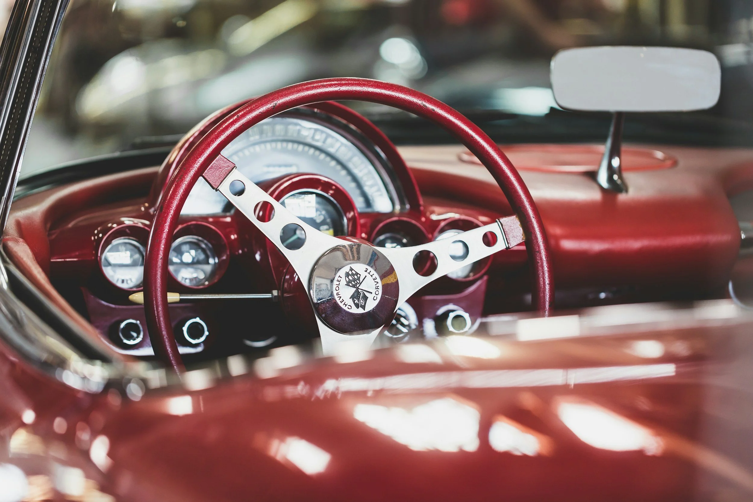 Interior of a vintage red Chevrolet Corvette sports car, showing the steering wheel with the Corvette logo, dashboard with gauges, and rearview mirror.