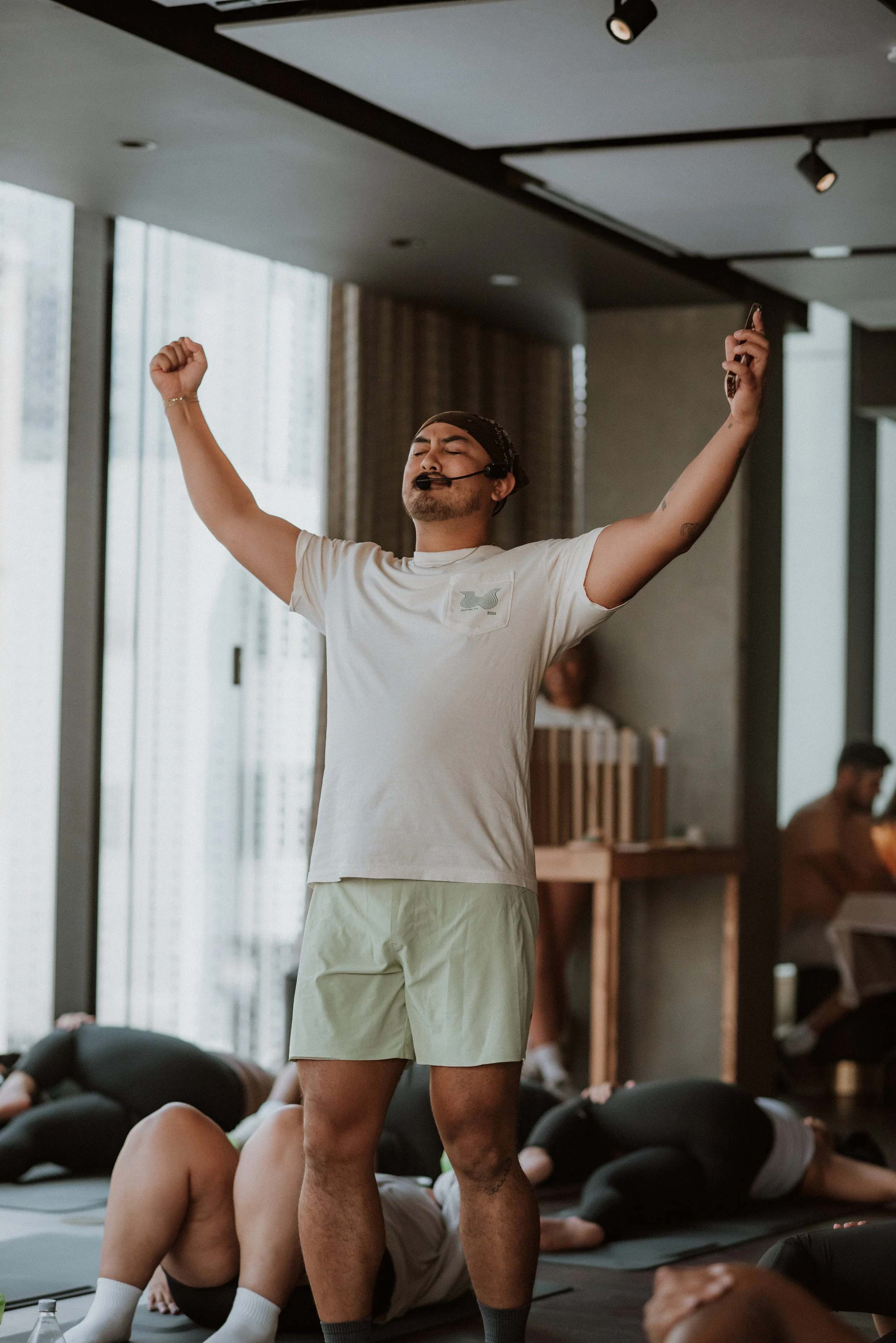 A man wearing a headset with a microphone, standing with arms raised in a yoga or exercise class, surrounded by other participants lying on mats in a bright indoor space.